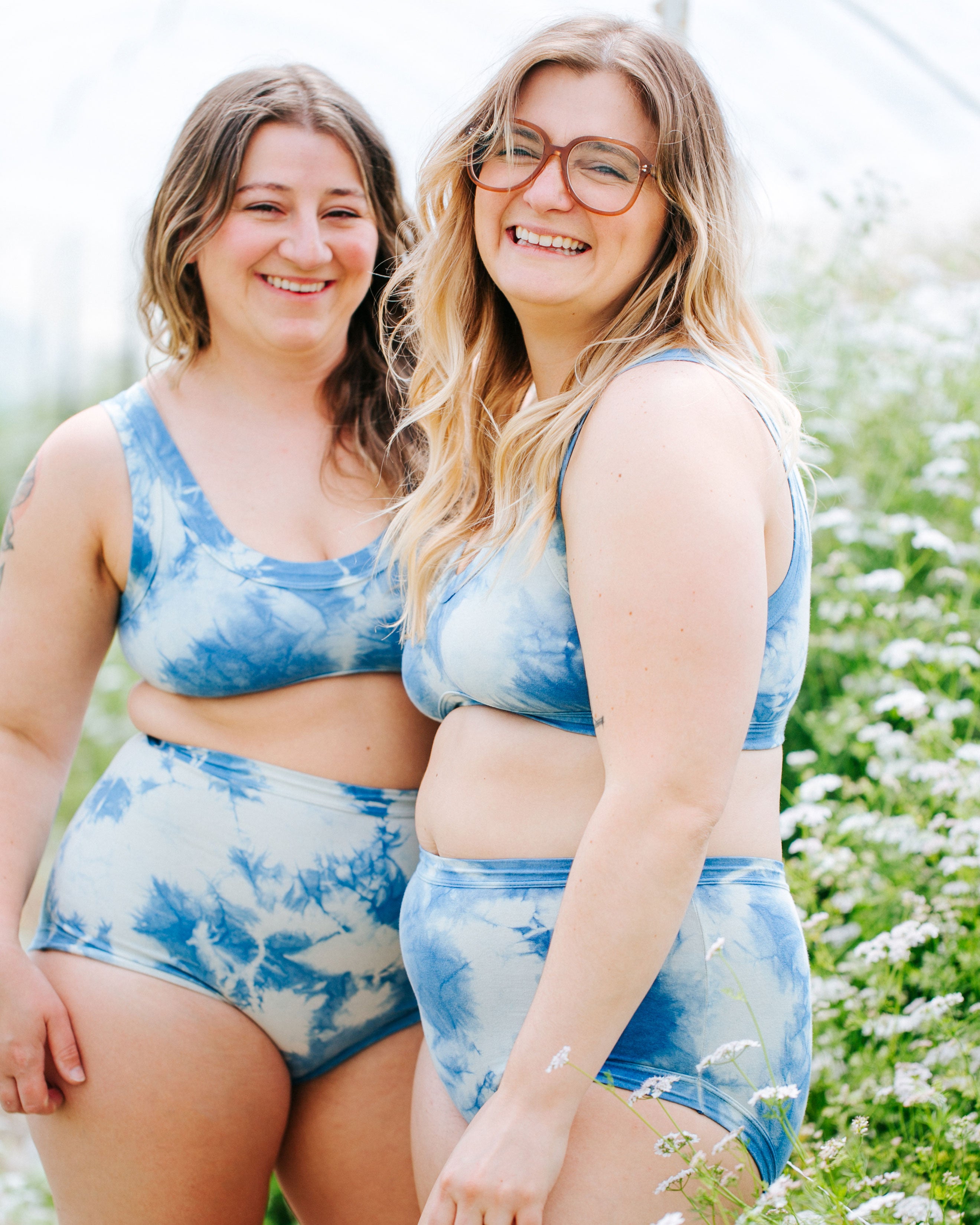 Two models smiling in a greenhouse wearing sets of indigo dyed Sauvie Skies sets of Sky Rise style underwear, Original style underwear, and Bralettes.