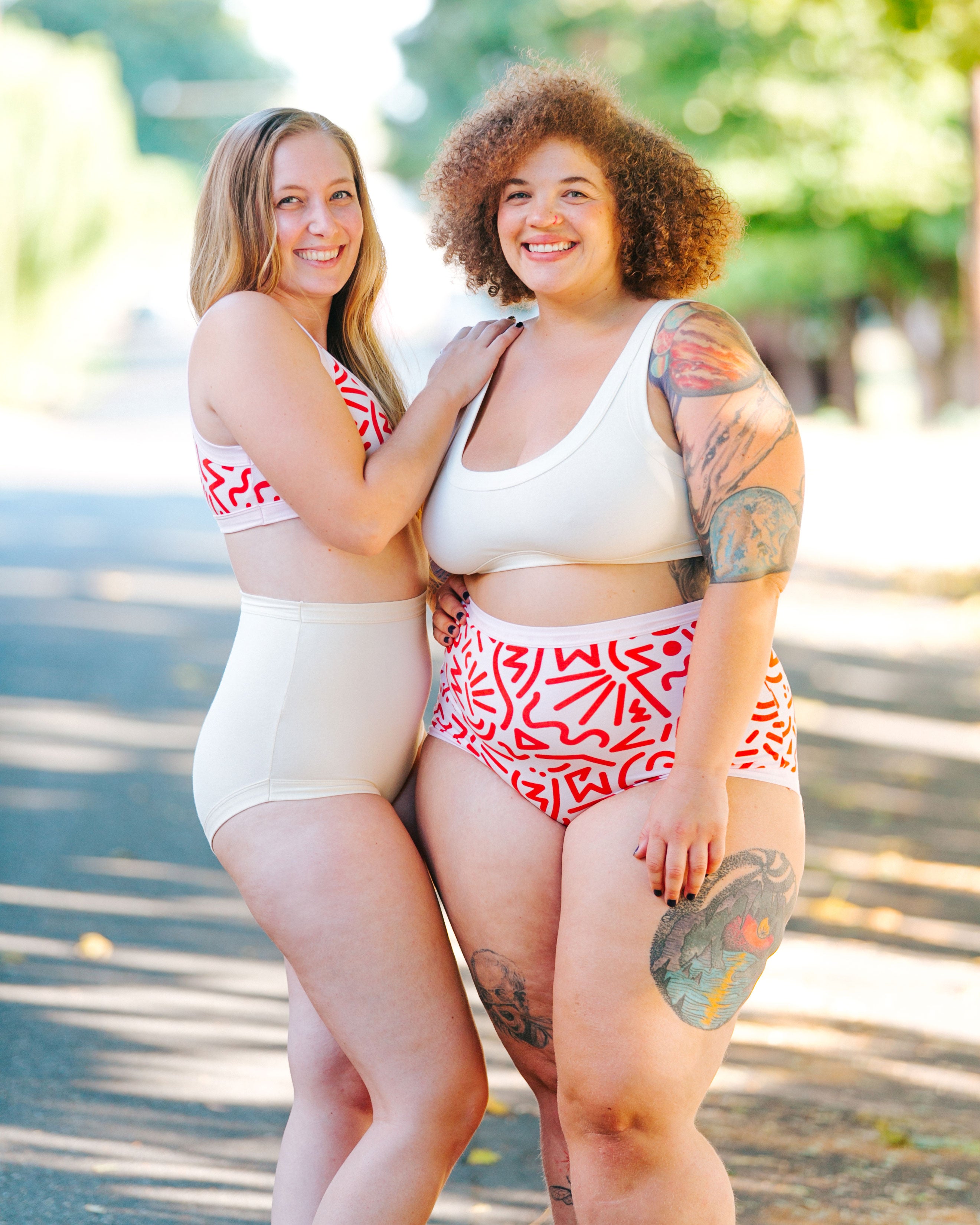 Two models standing together wearing Thunderpants organic cotton Sky Rise style underwear and Bralettes in both plain Vanilla and Energy Vibes print: large red squiggles printed on Perfect Pink.