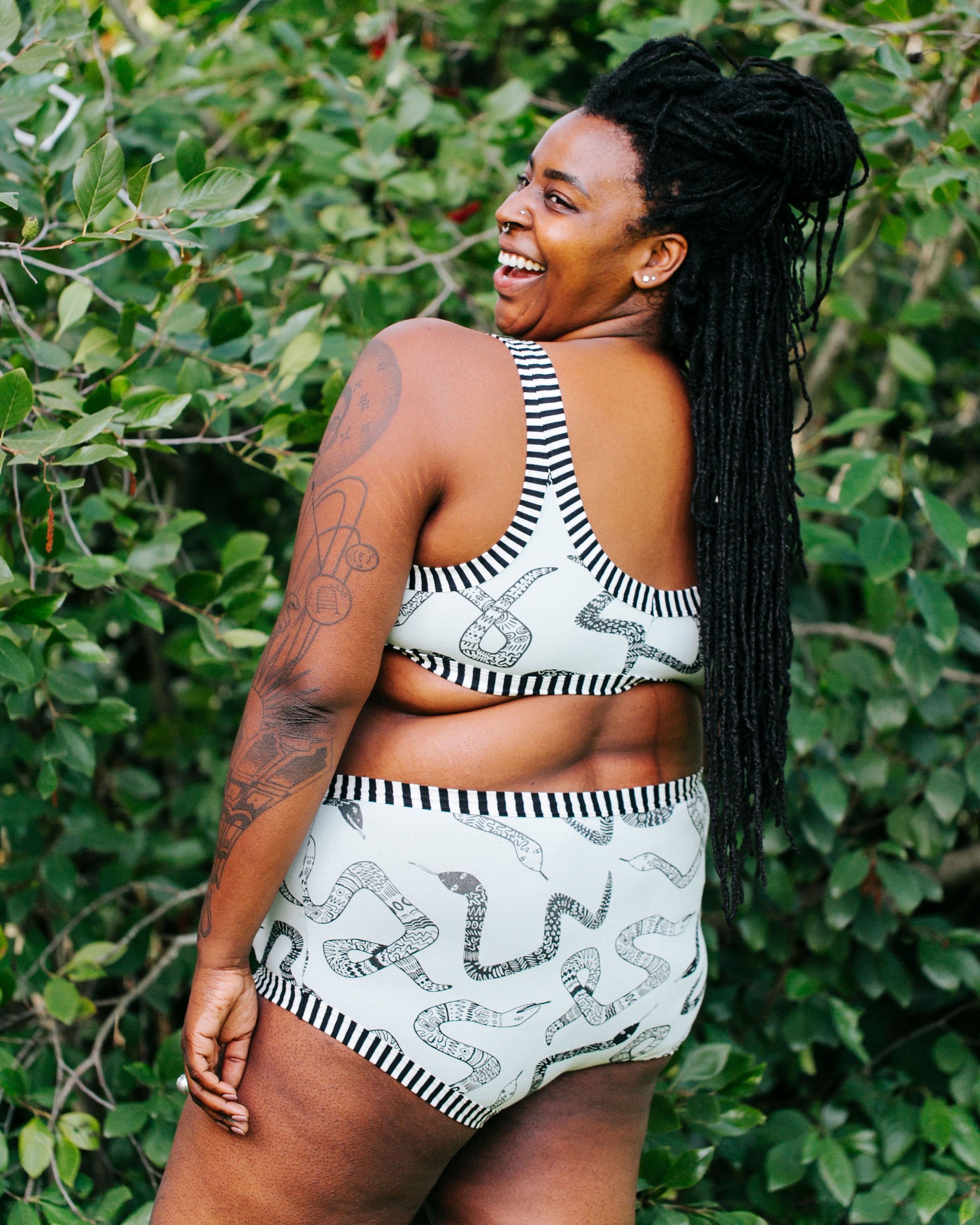 Model smiling over her shoulder with her back to us wearing Thunderpants organic cotton Bralette and Original style underwear in our Sketchy Snakes print: sage color with black sketched snakes and black and white stripe binding.