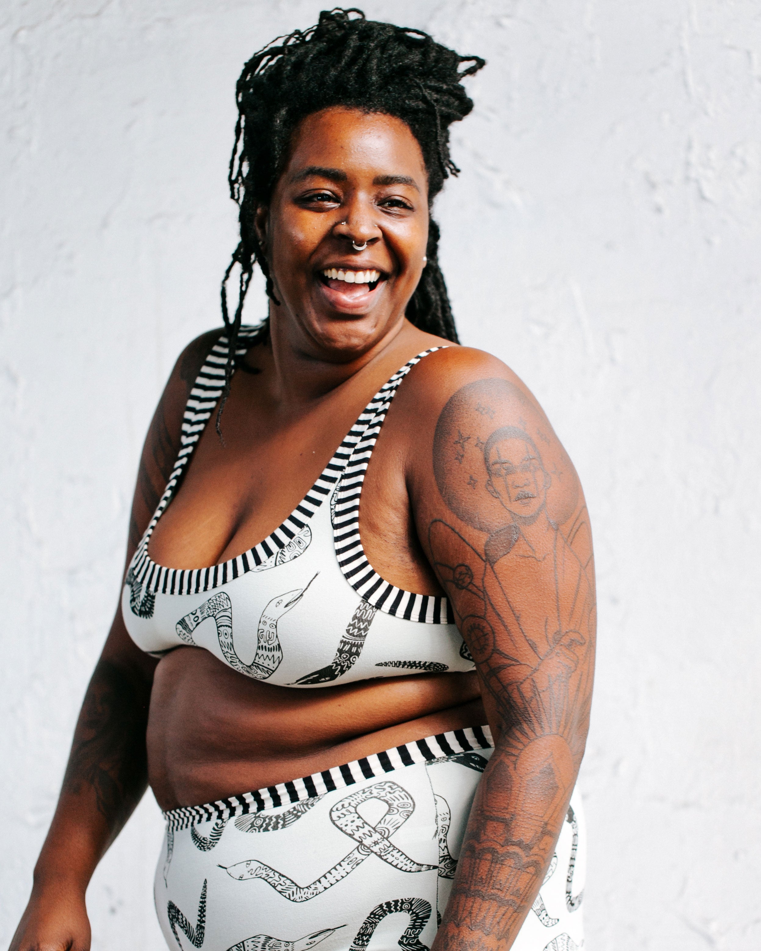 Model smiling at the camera while wearing Thunderpants organic cotton Bralette and Original style underwear in our Sketchy Snakes print: sage color with black sketched snakes and black and white stripe binding.