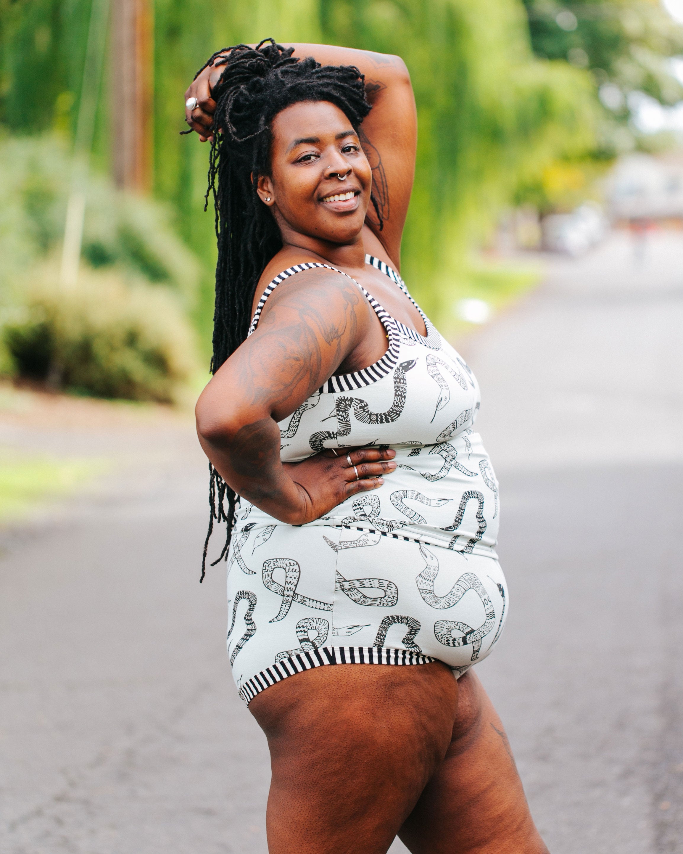 Model smiling at the camera outside wearing Thunderpants organic cotton Camisole and Original style underwear in our Sketchy Snakes print: sage color with black sketched snakes and black and white stripe binding.
