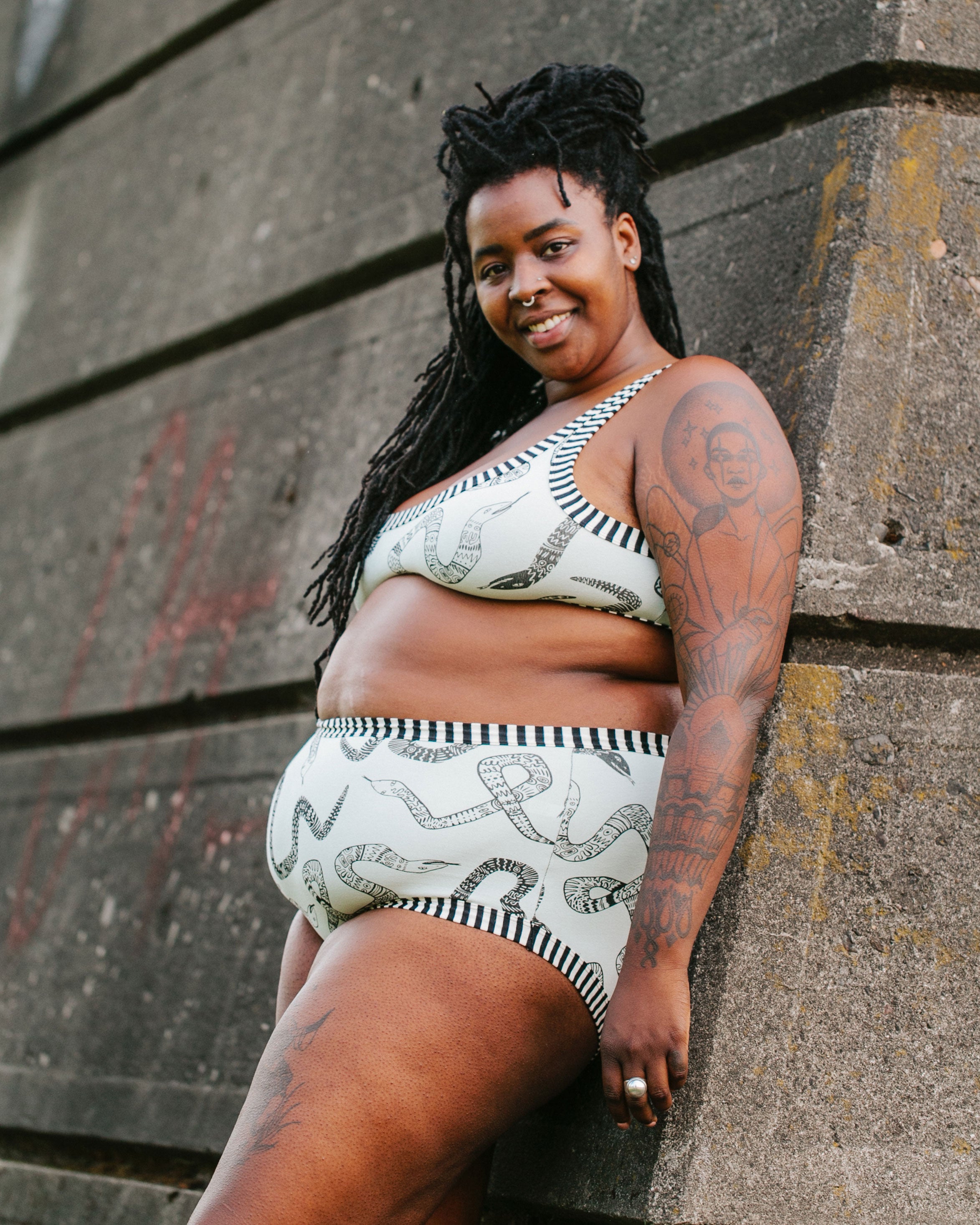 Model smiling outside wearing Thunderpants organic cotton Original style underwear and Bralette in our Sketchy Snakes print: sage color with black sketched snakes and black and white stripe binding.