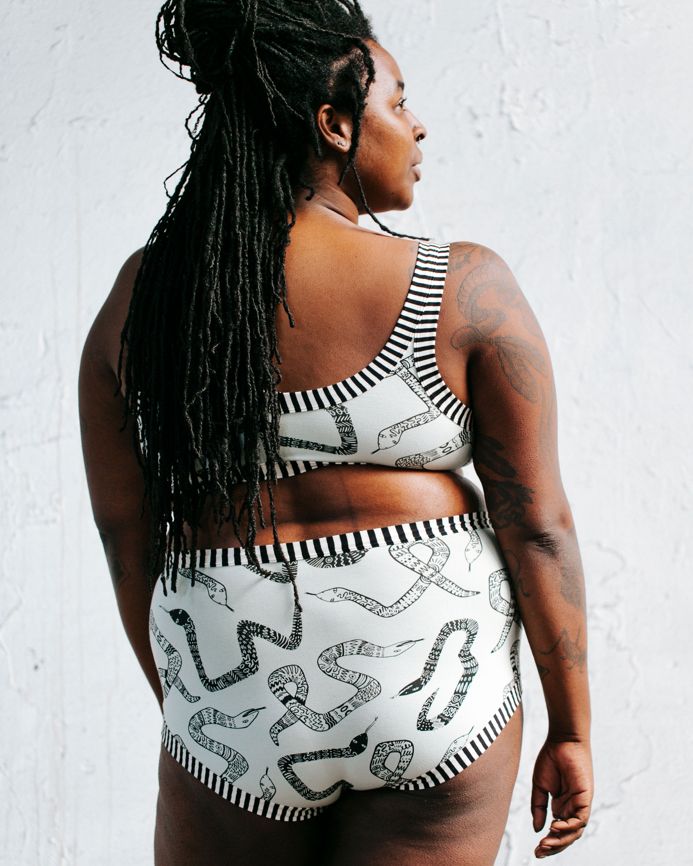 Model standing with her back to us wearing Thunderpants organic cotton Original style underwear and Bralette in our Sketchy Snakes print: sage color with black sketched snakes and black and white stripe binding.