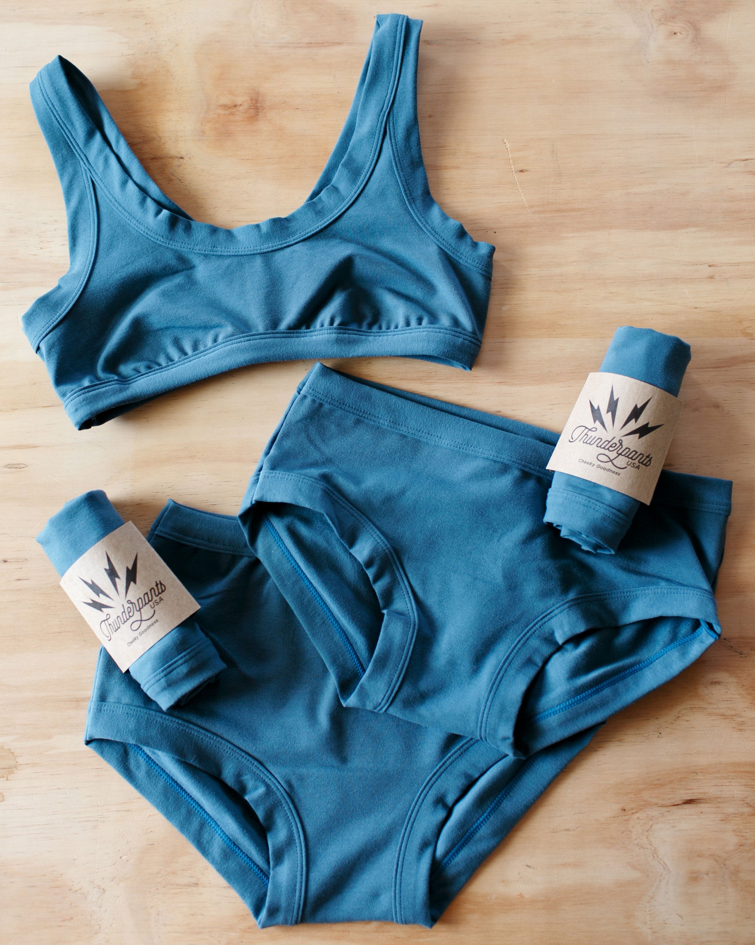 Flat lay of Thunderpants Organic Cotton Bralette, Hipster style and Original style underwear with packaged underwear in Stormy Blue color.
