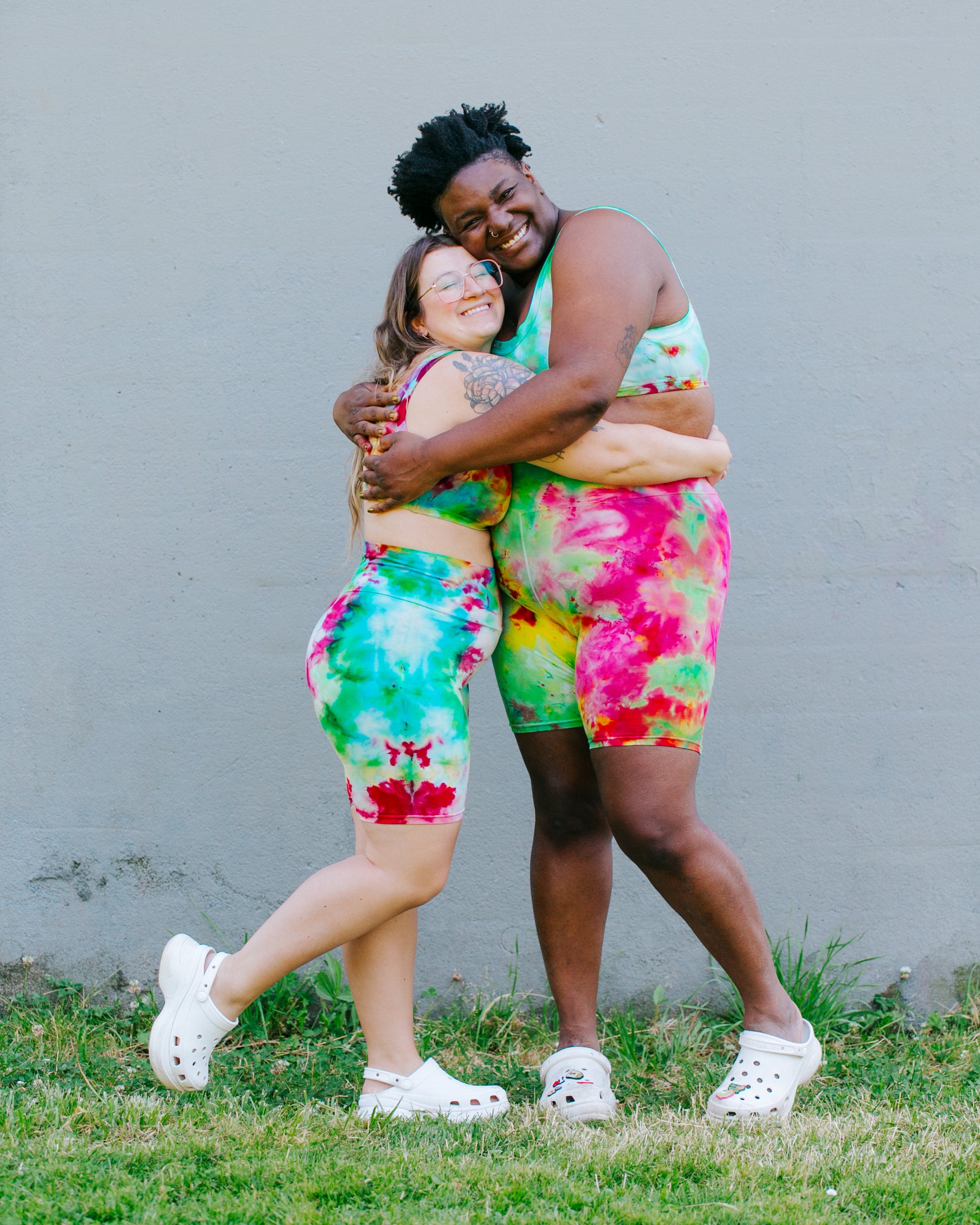 Two models smiling and hugging wearing sets of Ice Dyed Bike Shorts and Bralettes in a mix of green, pink, and yellow colors.