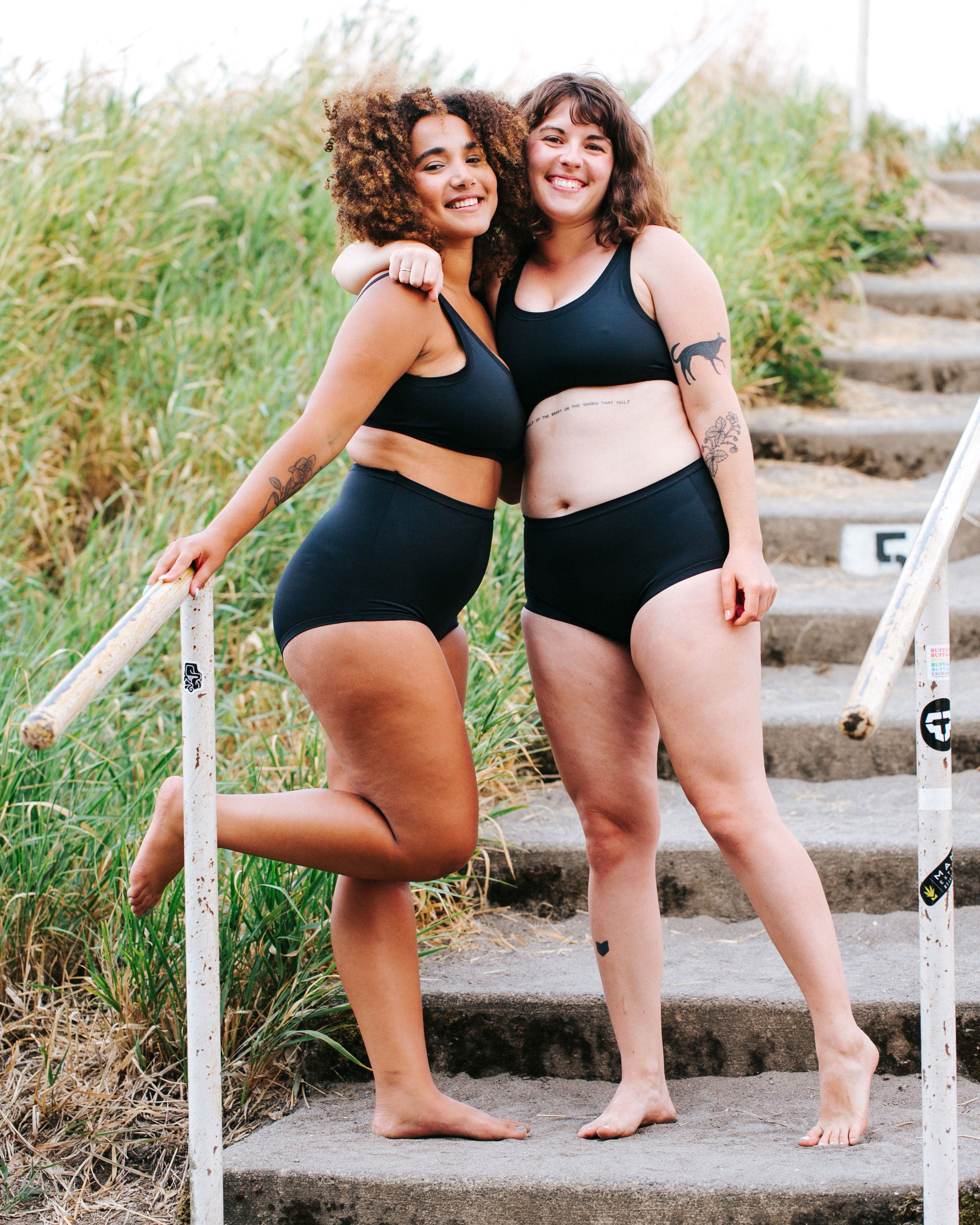 Two models hugging eachother and smiling wearing Thunderpants recycled nylon Sky Rise style and Original style Swimwear Bottoms and Swimwear Tops in Plain Black.