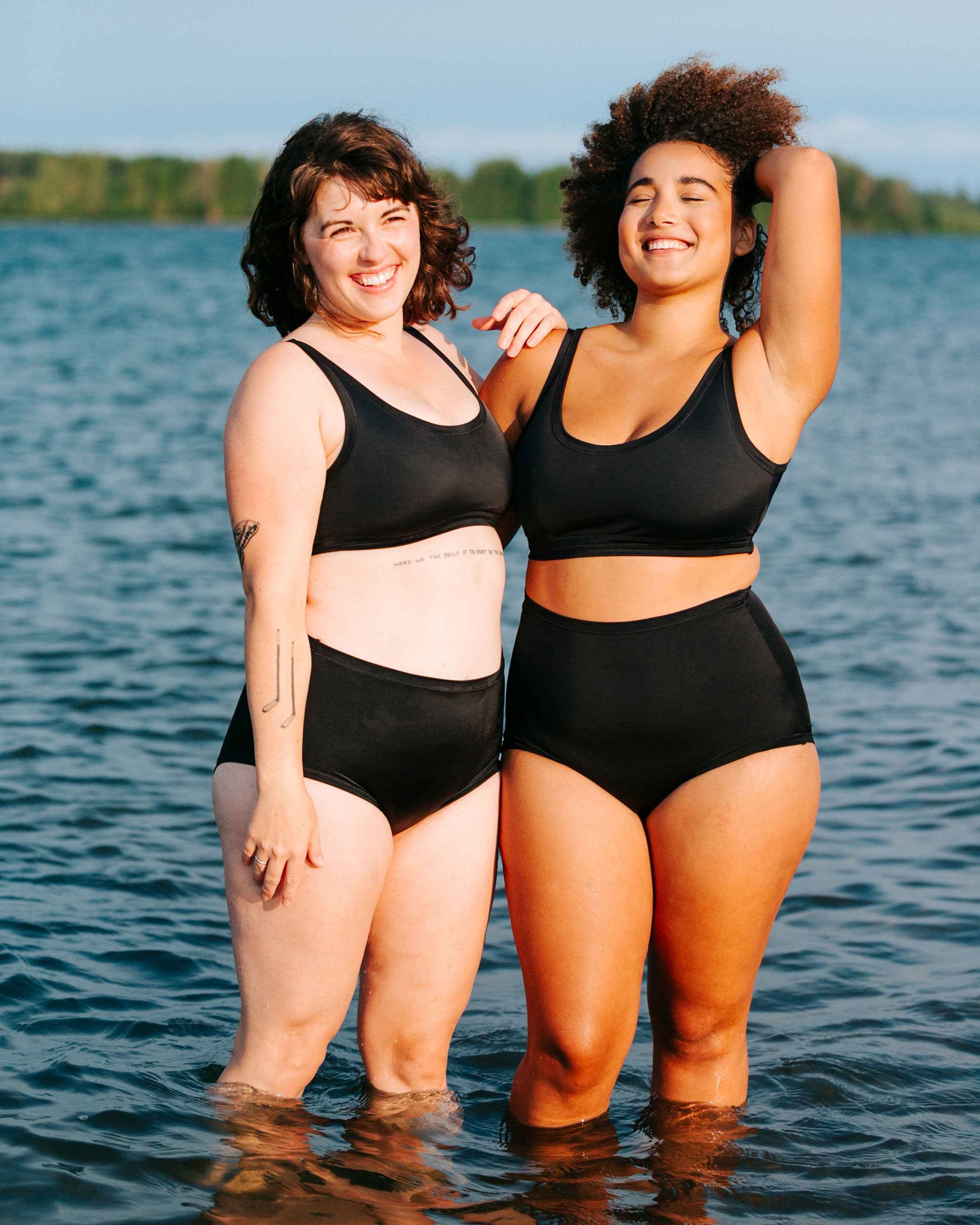 Two models standing together in the water wearing Thunderpants recycled nylon Original style and Sky Rise style Swimwear, and Swimwear Tops in Plain Black.