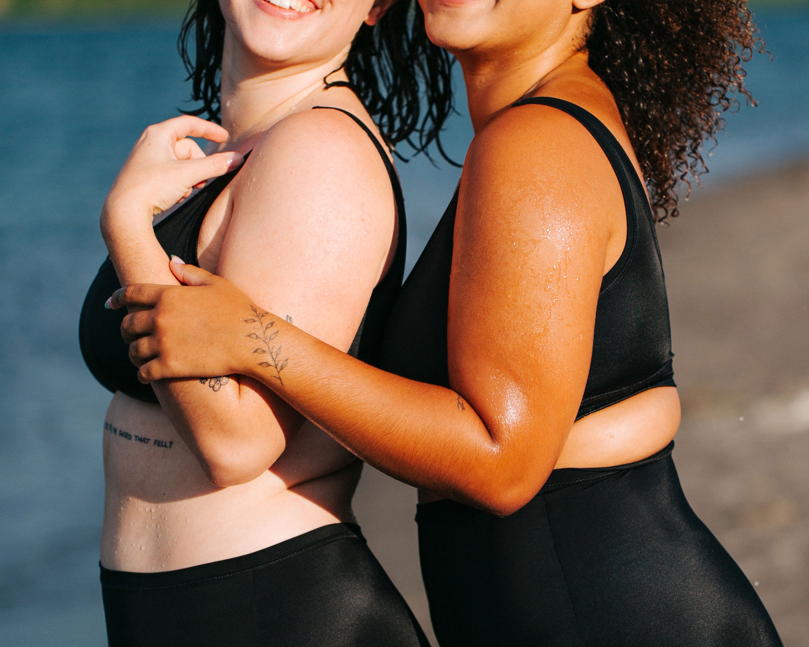 Two models from the side laughing together on the beach wearing Thunderpants recycled nylon Swimwear Original style Bottoms and Sky Rise style , and Swimwear Top in Plain Black.