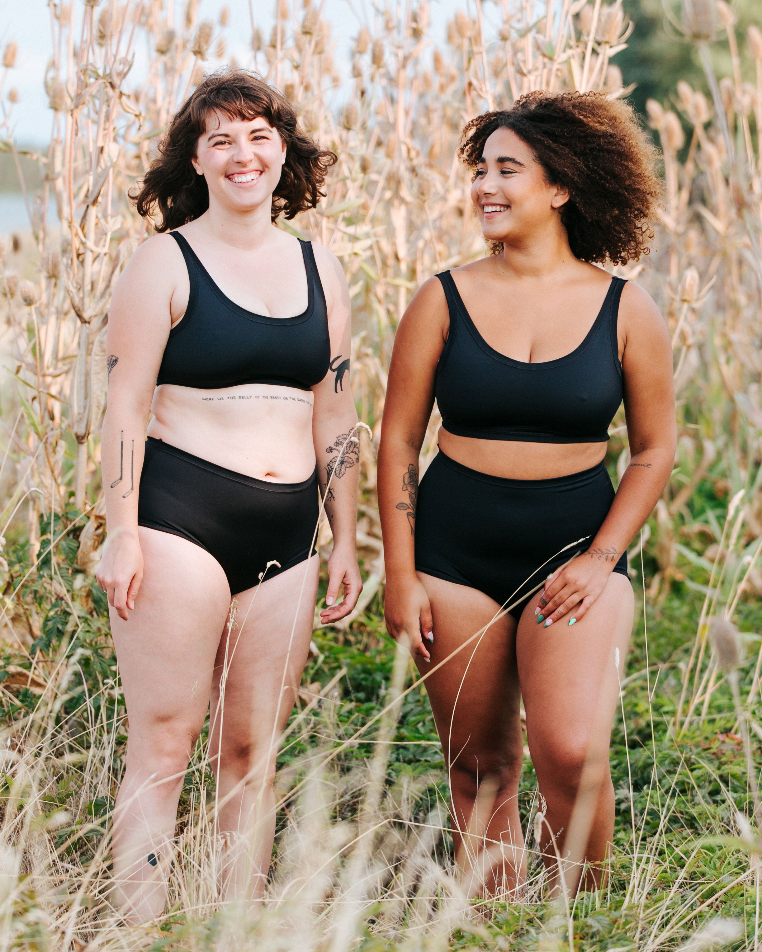 Two models laughing together on the beach wearing Thunderpants recycled nylon Swimwear Tops and Swimwear Original style and Sky rise style bottoms in Plain Black.