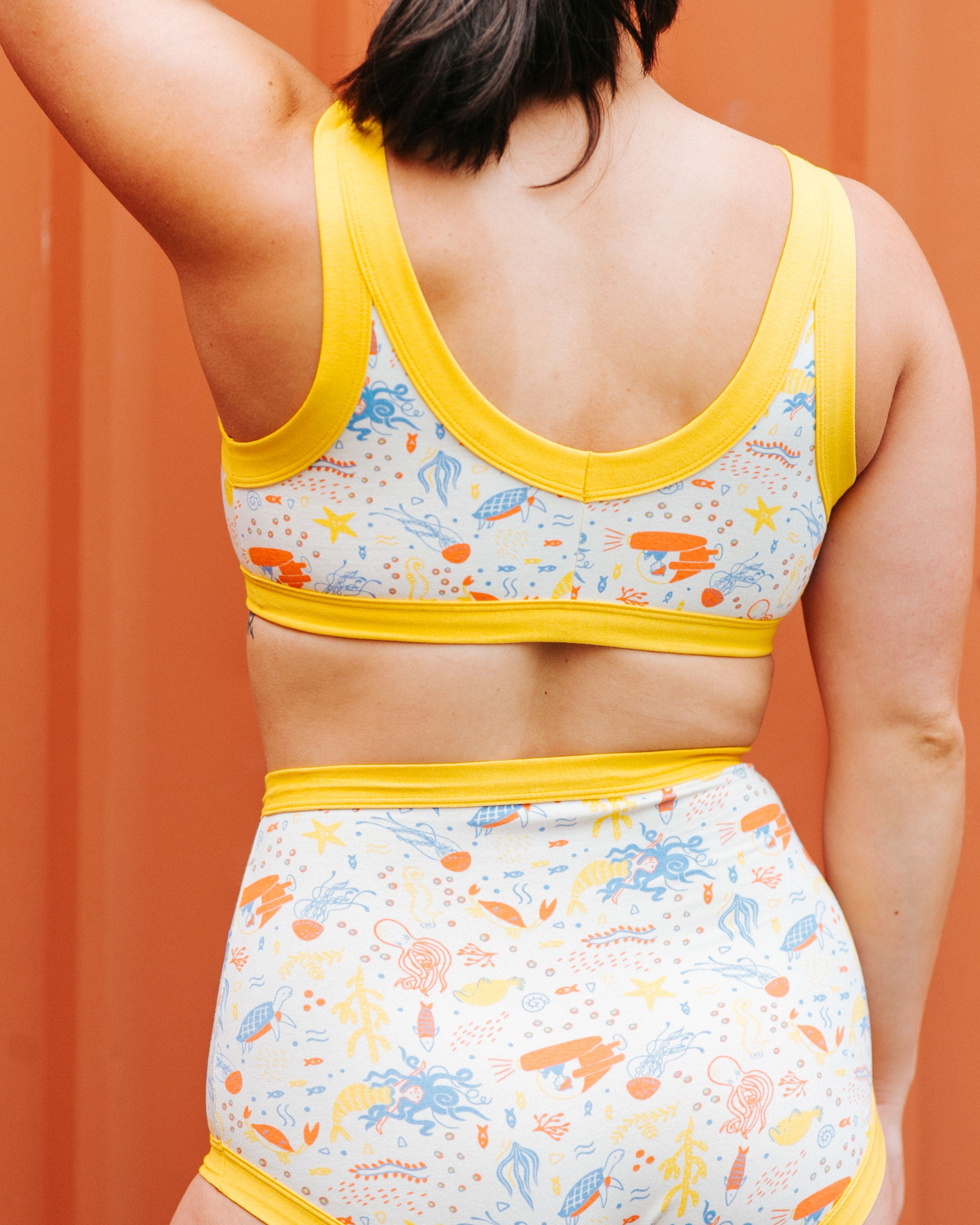 Close up photo of a model wearing Thunderpants organic cotton Bralette and Sky Rise style underwear in our Under the Sea print: Vanilla with small blue, orange, and gold underwater prints with Golden Yellow binding.