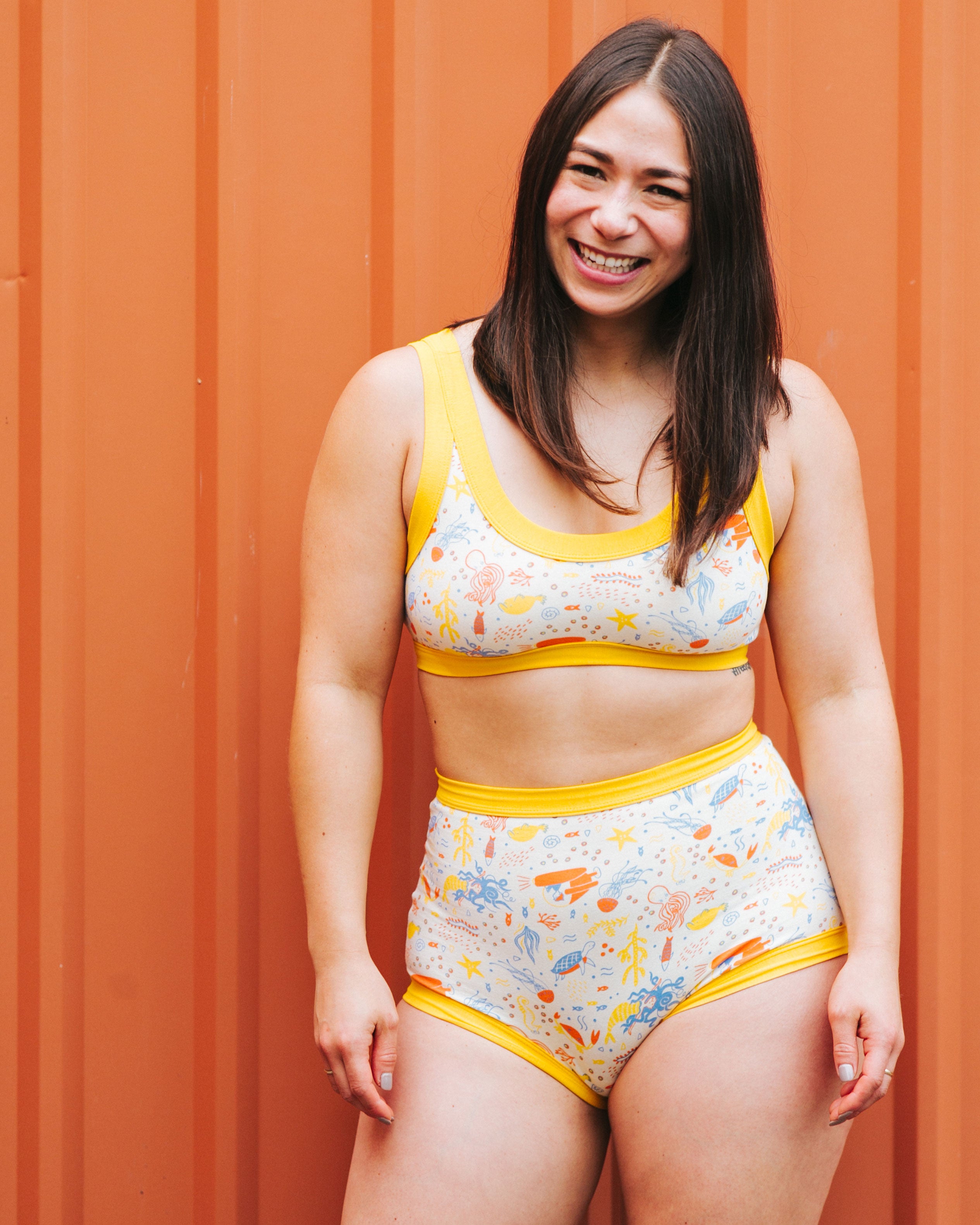 Model smiling wearing Thunderpants organic cotton Sky Rise style underwear and Bralette in our Under the Sea print: Vanilla with small blue, orange, and gold underwater prints with Golden Yellow binding.