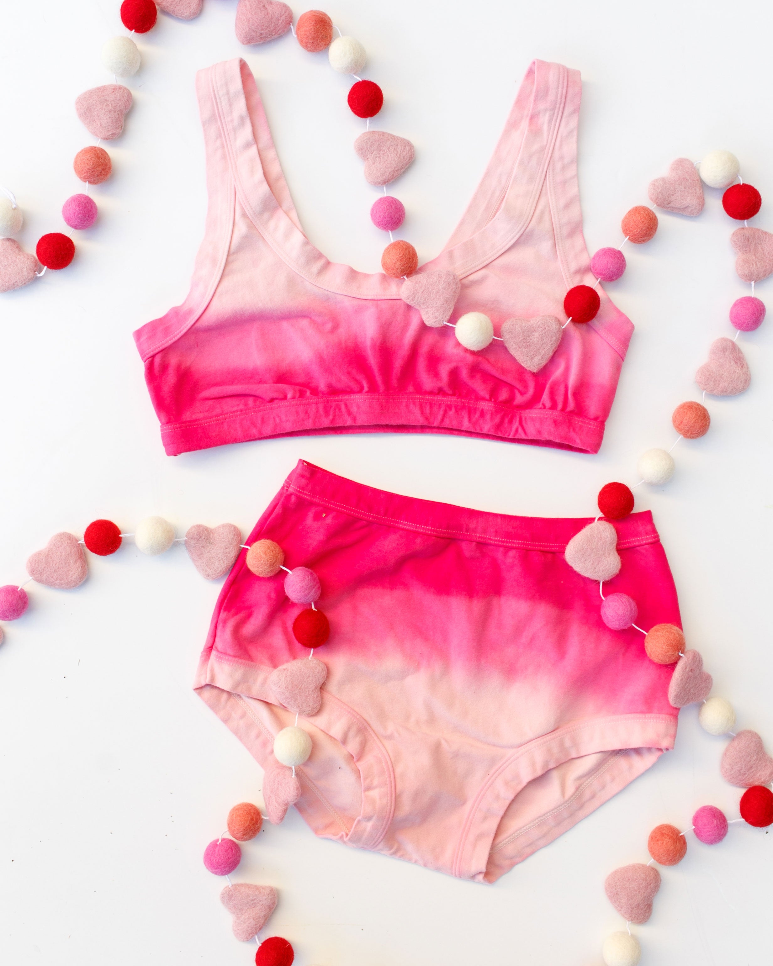 Flat lay of Thunderpants organic cotton Bralette and Original underwear in Valentine's Day Dip Dye ombre pinks with heart garland.