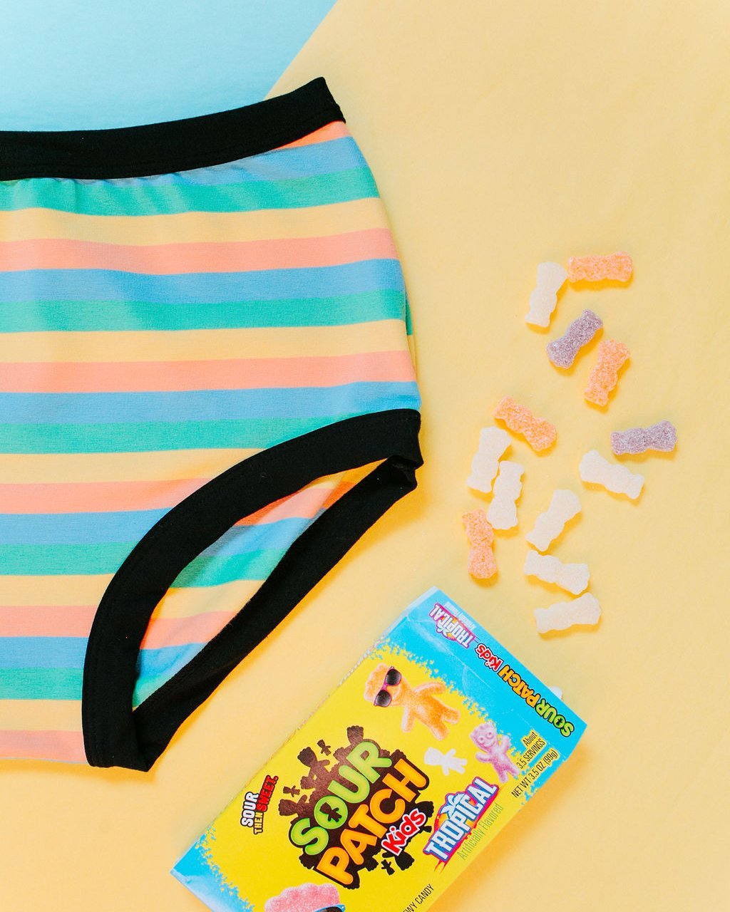 Flat lay of Thunderpants organic cotton Women’s Original style underwear in Pastel Rainbow Stripes with bright blue and yellow background and sour patch candy.